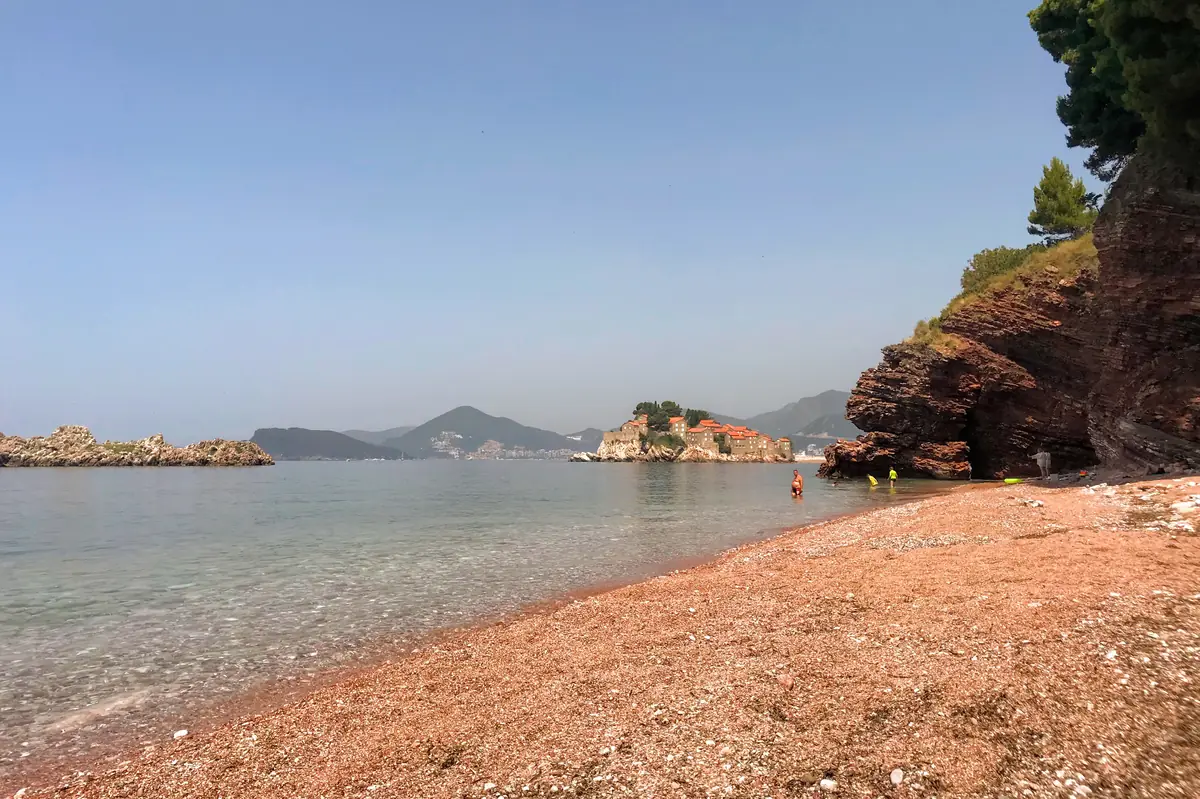 Crvena Stijena Beach near Sveti Stefan, showing the peaceful pebble shore, red cliffs, pine forests, and Sveti Stefan Resort in the background