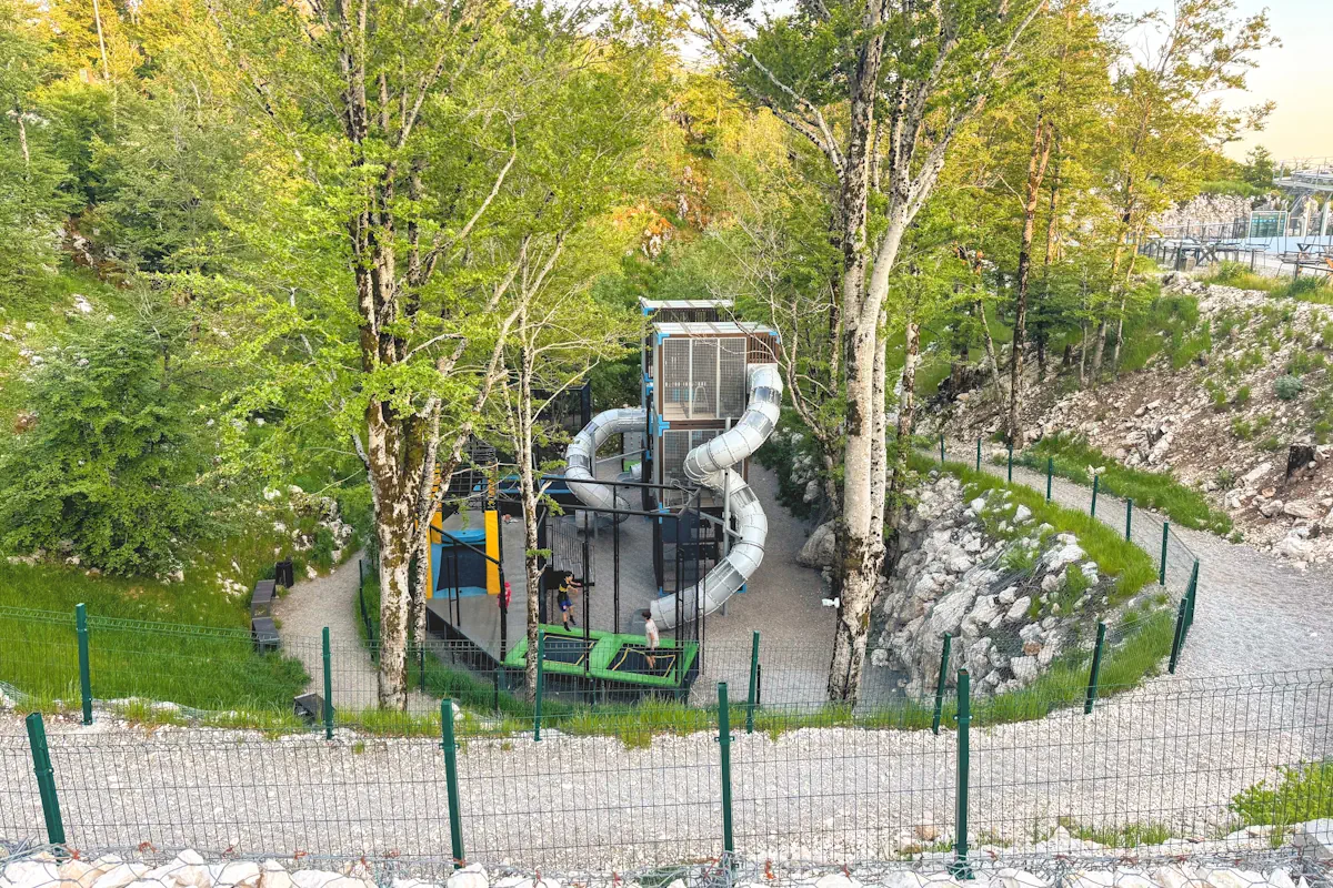 The children's adventure playpark at Kuk Station featuring a large multi-story tower slide, outdoor trampolines, and secure walking paths enclosed by green fencing in a forested alpine setting