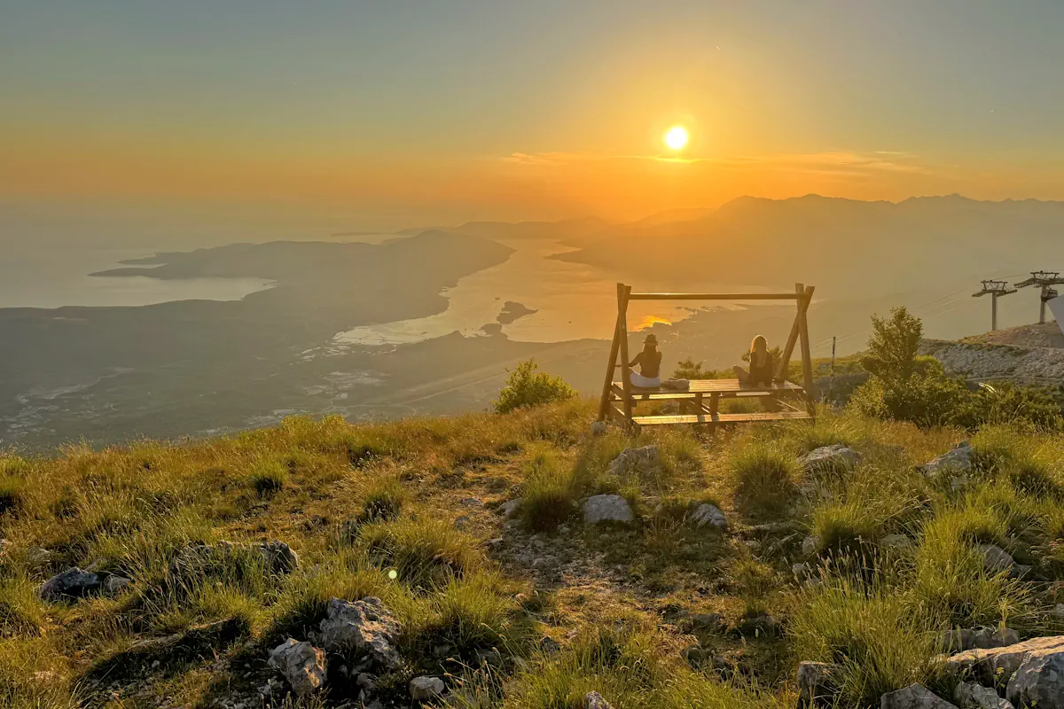 Two friends sitting quietly together on the rustic mountain sunset bench looking down at the sun dipping over the blue waters of Boka Bay and the distant Adriatic horizon