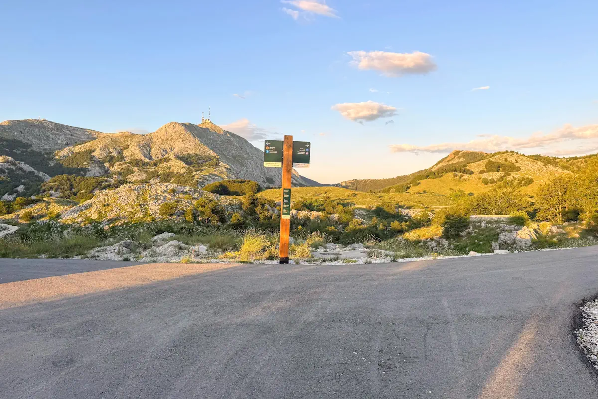 A spectacular ultra-wide mountain panorama looking over the limestone peaks of the Lovćen massif with an official wooden trail directory signpost in the center