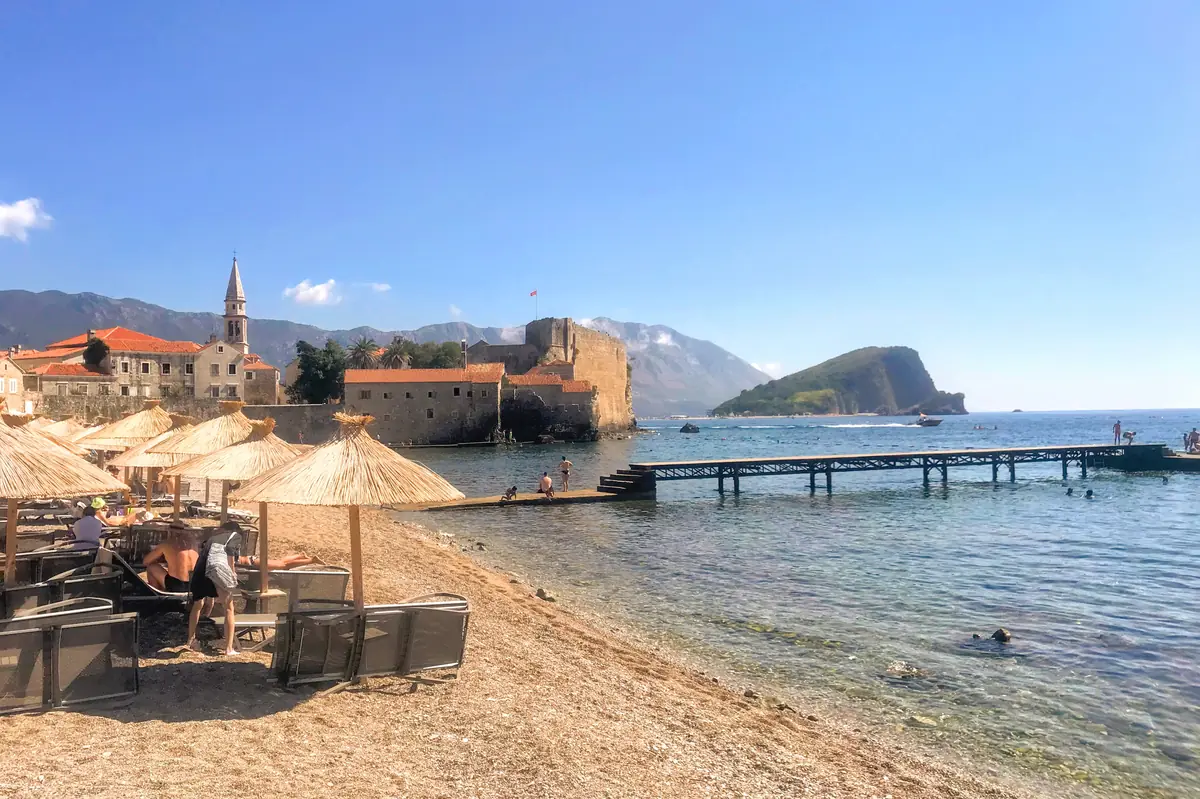 Ricardova Glava beach in Budva with people relaxing on sunbeds against the backdrop of Sveti Nikola Island and the ancient stone walls with bell tower