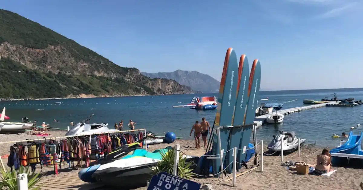 Wide view of Jaz Beach near Budva showing watersports rentals, towering mountains, and turquoise water