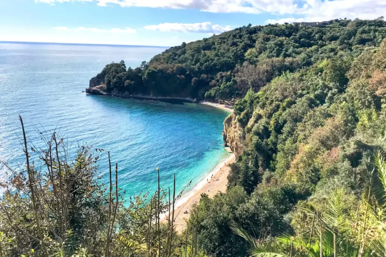 Aerial view of Mogren Beach in Budva, Montenegro, showing the golden pebble shoreline, turquoise Adriatic water, and the dramatic sandstone cliffs separating Mogren I and II