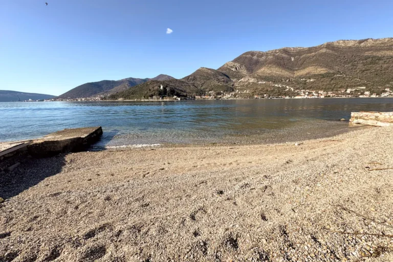 A narrow pebble beach at Opatovo on the Tivat Riviera, showing clear Adriatic water and the coastal road stone wall under a bright sky.