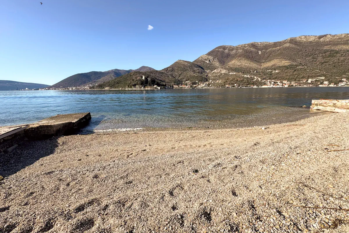 A narrow pebble beach at Opatovo on the Tivat Riviera, showing clear Adriatic water and the coastal road stone wall under a bright sky.