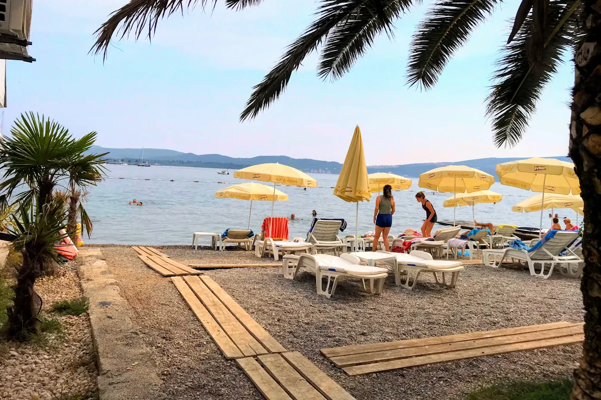 The pebble shoreline and turquoise water of Pona Seljanovo Beach in Tivat, with palms and sunbeds overlooking the Porto Montenegro.